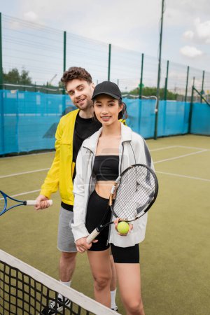 Photo for Cheerful man and woman in active wear holding tennis rackets and ball on court, looking at camera - Royalty Free Image