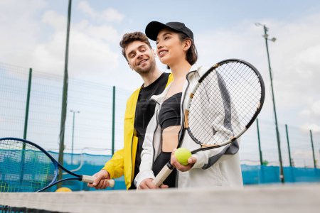 Photo for Cheerful couple in stylish active wear holding tennis rackets and ball on court, lifestyle and sport - Royalty Free Image