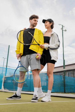 happy woman in cap and active wear holding racket and ball while hugging boyfriend on court, sport