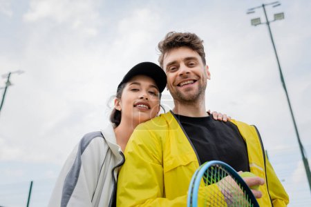Foto de Mujer feliz en gorra y desgaste activo abrazo novio sosteniendo raqueta en la cancha, deporte, vista de bajo ángulo - Imagen libre de derechos