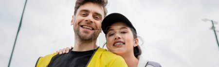 banner, happy woman in cap and active wear hugging boyfriend on tennis court, sport, low angle view