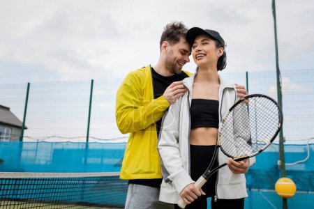 glücklicher Mann in aktiver Kleidung umarmt Freundin in Mütze mit Tennisschläger auf dem Platz, Lifestyle und Sport