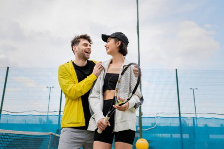 positive man in active wear hugging woman in cap with tennis racket on court, lifestyle and sport