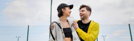 Foto de Pancarta, hombre positivo en desgaste activo abrazar a la mujer en la gorra con raqueta de tenis en la cancha, estilo de vida - Imagen libre de derechos