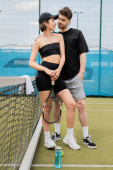 positive man in black active wear hugging woman in cap with tennis racket on court, lifestyle Stickers #665315744