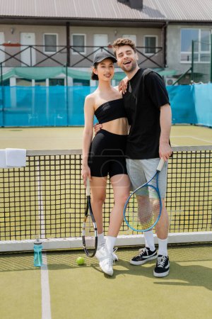 romance on tennis court, positive couple in black active wear standing with tennis rackets near ball magic mug #665315764