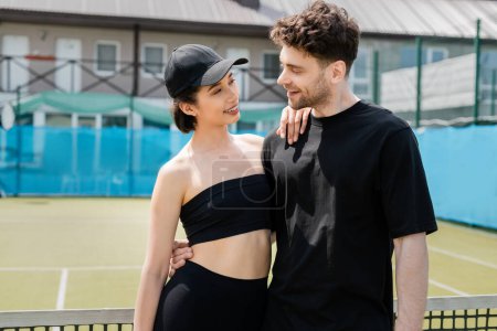 positive couple in black active wear standing near tennis net, romance on tennis court