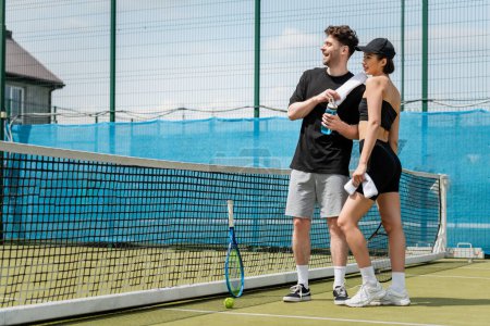 Foto de Feliz pareja de pie con toalla y botella deportiva cerca de la red de tenis, raqueta y pelota en la cancha - Imagen libre de derechos