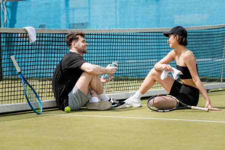 heureux couple assis avec serviette et bouteille de sport près du filet de tennis, raquette et balle sur le court