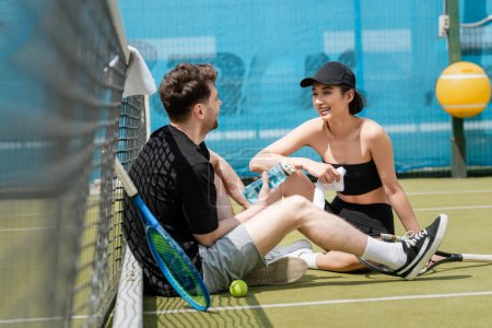 Foto de Feliz pareja en desgaste activo sentado con botella deportiva cerca de la red de tenis, raqueta de tenis y pelota, - Imagen libre de derechos
