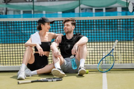 Photo for Happy couple resting on tennis court, sitting together near tennis net, sports bottle, rackets, ball - Royalty Free Image