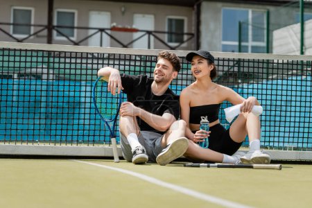 Foto de Hombre y mujer felices en ropa deportiva descansando cerca de la red de tenis con raquetas en la cancha, estilo de vida saludable - Imagen libre de derechos