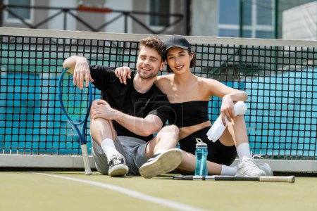 Photo for Happy man and woman in sportswear resting near tennis net on court, ball, rackets, looking at camera - Royalty Free Image