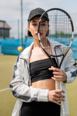 sporty woman in active wear and cap looking at camera through tennis racket on court, sport hoodie #665316032