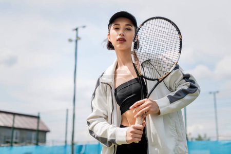 sportliche Frau in aktiver Kleidung und Mütze blickt in die Kamera und hält Tennisschläger auf dem Platz, Sport