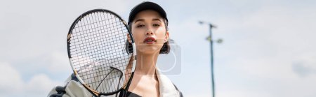 bannière, femme en tenue active et casquette regardant la caméra et tenant une raquette de tennis sur le court, sport