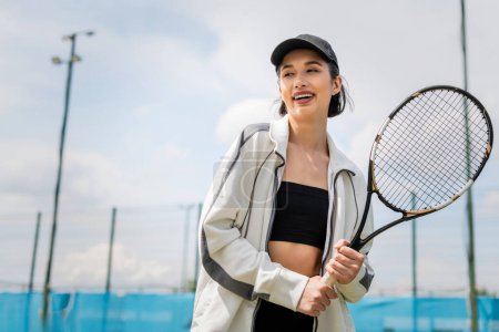 szczęśliwa kobieta w aktywnym ubraniu i czapce z rakietką tenisową na korcie, tenisistka, motywacja