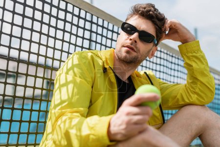 athletischer Tennisspieler mit Sonnenbrille, der neben dem Tennisnetz sitzt und Ball, Sport und Stil in der Hand hält