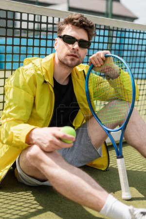 przystojny mężczyzna w okularach przeciwsłonecznych przy siatce tenisowej, trzymając rakietę i piłkę, sport i styl, aktywny strój