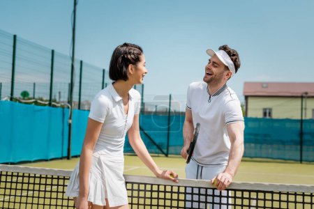 happy man looking at woman near tennis net, cheerful couple standing on tennis court, active wear