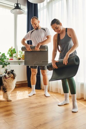 Cheerful couple in sportswear holding fitness mats near border collie in living room at home