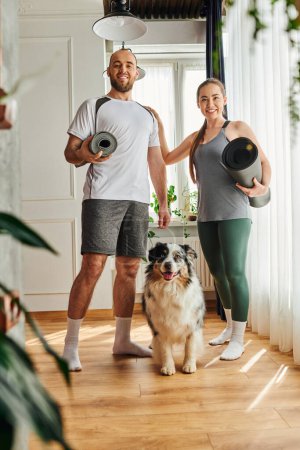 Smiling couple in sportswear holding fitness mats and looking at camera near border collie at home