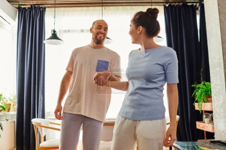 Smiling bearded man in homewear holding hand of girlfriend while spenging time together at home