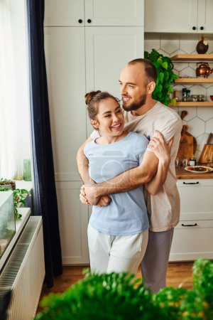 Foto de Sonriente hombre en ropa de casa abrazando novia morena mientras está de pie cerca de la ventana en la cocina en casa - Imagen libre de derechos