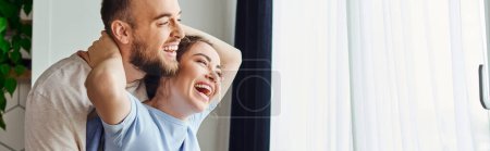 Positive woman hugging boyfriend in homewear while standing together near window at home,banner