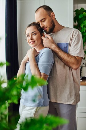 Tender bearded man touching smiling girlfriend in homewear while spending time together at home