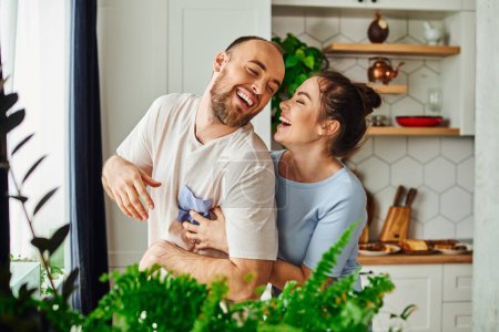 Joyful brunette woman in loungewear embracing bearded boyfriend near green plants at home