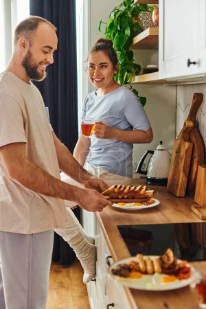 Mujer sonriente sosteniendo el té mientras que el novio en la ropa de casa tomar platos con desayuno en la cocina