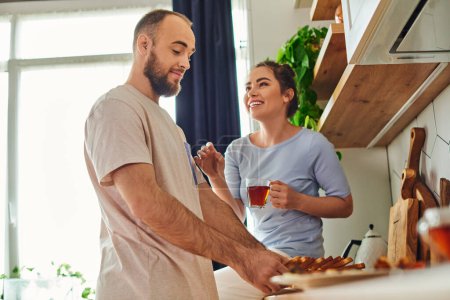 Smiling woman in homewear holding tea and talking to boyfriend taking bread during breakfast at home