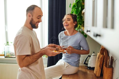 Fröhlicher Mann in Homewear hält Teller mit leckerem Frühstück bei Freundin mit Tee zu Hause