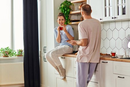 Foto de Mujer sonriente en ropa de casa sosteniendo té cerca de novio con desayuno en el plato en la cocina por la mañana - Imagen libre de derechos