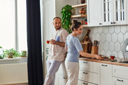 Lächelnder Mann in Homewear hält Tee, während Freundin morgens in der Küche frühstückt