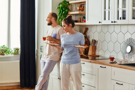 Smiling couple in homewear holding tea and tasty breakfast in kitchen in morning at home