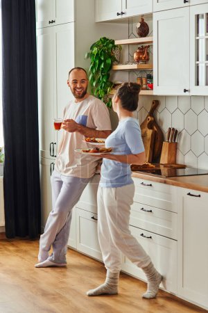Cheerful bearded man in homewear holding tea and looking at girlfriend with preakfast in kitchen