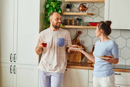 Foto de Hombre positivo sosteniendo el té y tomando el plato con el desayuno de su novia mientras está de pie en la cocina - Imagen libre de derechos