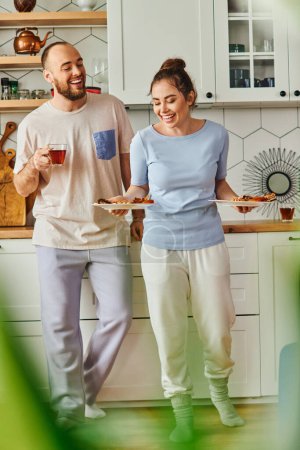 Smiling couple in homewear holding tasty breakfast on plates and tea in morning in kitchen at home