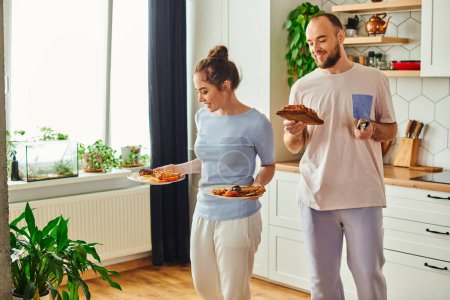 Smiling couple in homewear holding toasts and plates with tasty breakfast at home in morning