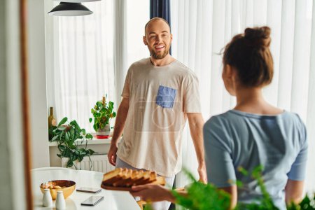 Smiling man in homewear standing near blurred girlfriend with homemade breakfast in morning at home