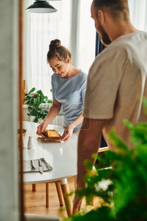 Smiling woman in homewear putting toasts on table near blurred boyfriend at home in morning