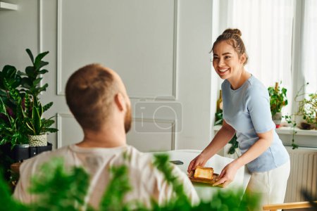 Fröhliche Frau in Hauskleidung stellt Toasts auf den Tisch neben verschwommenem Freund beim Frühstück zu Hause