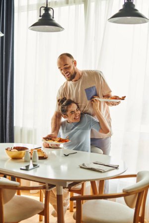 Positive woman in homewear sitting near boyfriend putting breakfast on table at home in morning