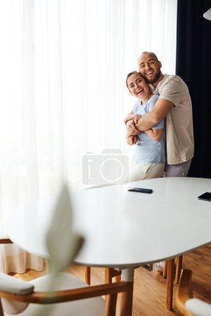 Lächelndes Paar in Homewear umarmt sich und blickt in die Kamera neben Smartphones auf dem heimischen Tisch