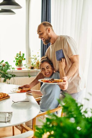 Positive brunette woman touching boyfriend putting tasty breakfast on table at home in morning