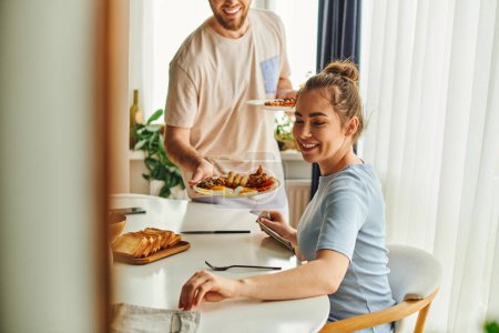 Photo for Smiling woman sitting near cutlery and blurred boyfriend holding tasty breakfast at home in morning - Royalty Free Image