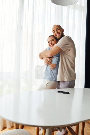 Smiling bearded man in homewear hugging girlfriend near smartphone on table at home