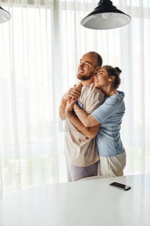 Mujer sonriente en ropa de casa abrazando novio y de pie cerca de teléfono inteligente con pantalla en blanco en casa